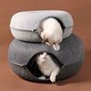 Two fluffy white cats are peeking out of the tunnel hideaway of a gray felt cat bed.