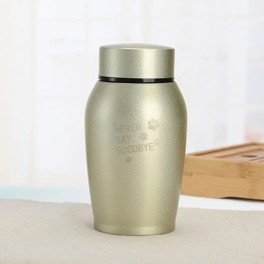 A matte gold stainless steel pet memorial storage box with a black lid sitting on a light surface.