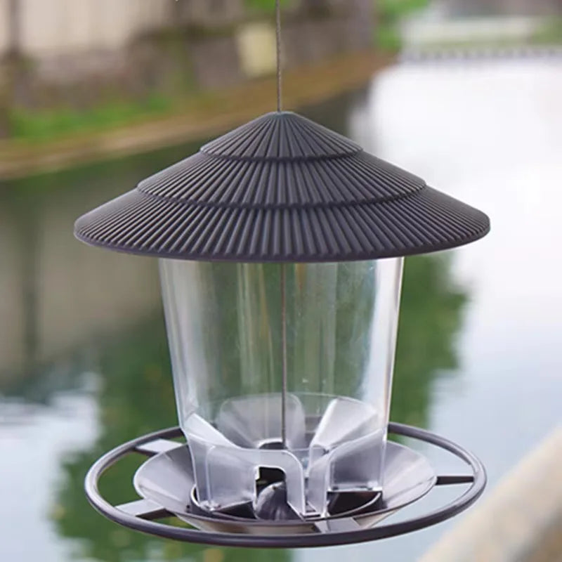Wasserfester Vogelfuttermittelspender mit klarem Glaskörper und dunklem grauen Kegeldach, über Wasser hängend.