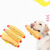 A light-colored dog is holding a large yellow hot dog squeaky chew toy with red ends.