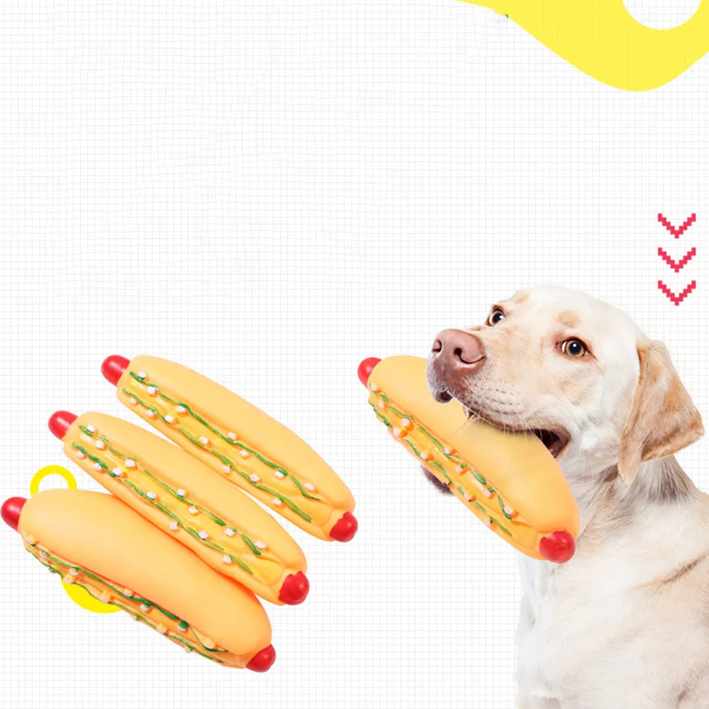 A light-colored dog is holding a large yellow hot dog squeaky chew toy with red ends.