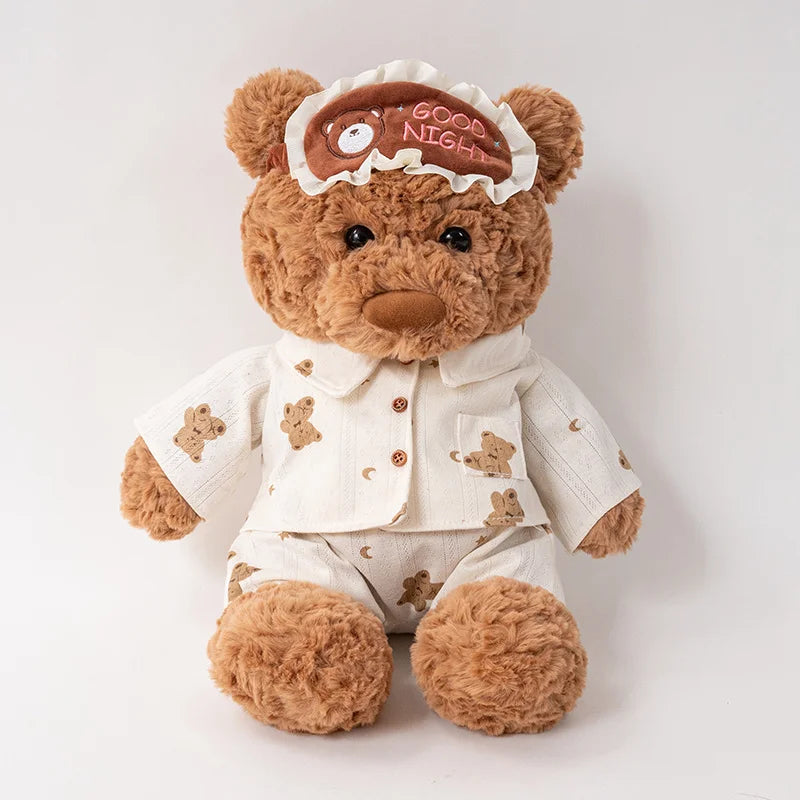 A brown teddy bear plush wearing a light-colored outfit and an eye mask on its head.