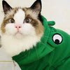 Eine Katze mit einem grünen Frosch-Hoodie und schwarzem Augenabdeckung, die direkt in die Kamera schaut.