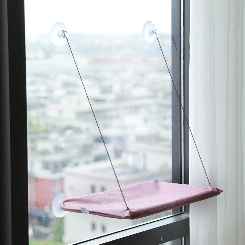 A pink hanging hammock product suspended by thin cords in a window frame overlooking a city skyline.