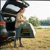 Eine Frau öffnet die Motorhaube eines Autos und hält eine grüne portable Katzenhütte draußen.