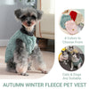 A small, fluffy dog wearing a teal fleece vest in an autumn winter setting.