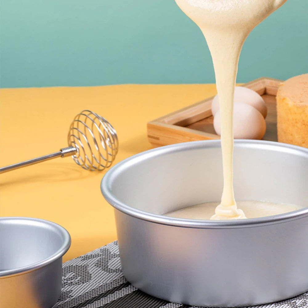 A stream of light beige batter pours into a silver round aluminum cake pan, set against a yellow background with eggs and a w