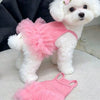A white poodle wearing a pink tutu dress, with a separate pink tutu lying nearby.