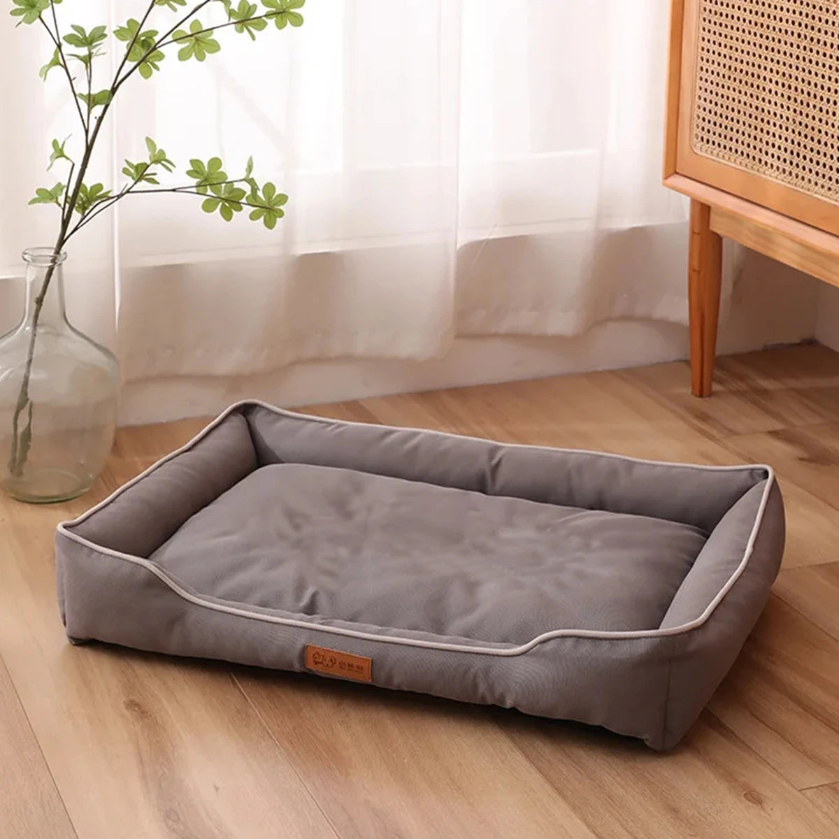 A gray waterproof orthopedic dog bed is placed on a wooden floor next to a window with greenery.