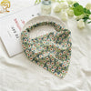 A floral patterned triangle head scarf is displayed on a white cloth next to a glass bottle and a piece of paper.