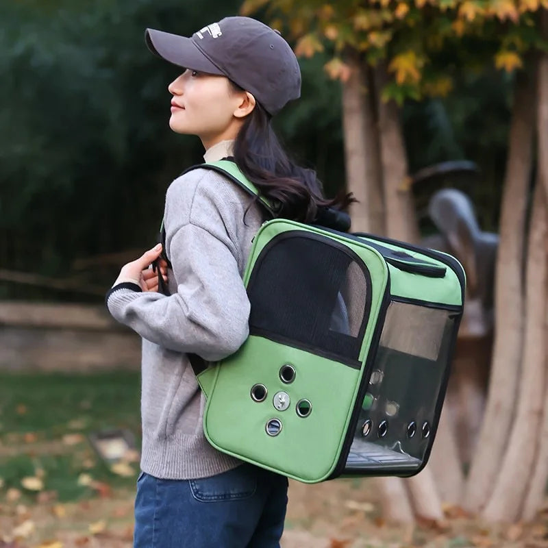 A woman wearing a grey sweater and cap holds a green ventilated bird travel backpack with a perch and feeding cups outdoors.