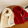 A beige knitted scarf with a cat embroidery patch lies next to a red knitted sweater.