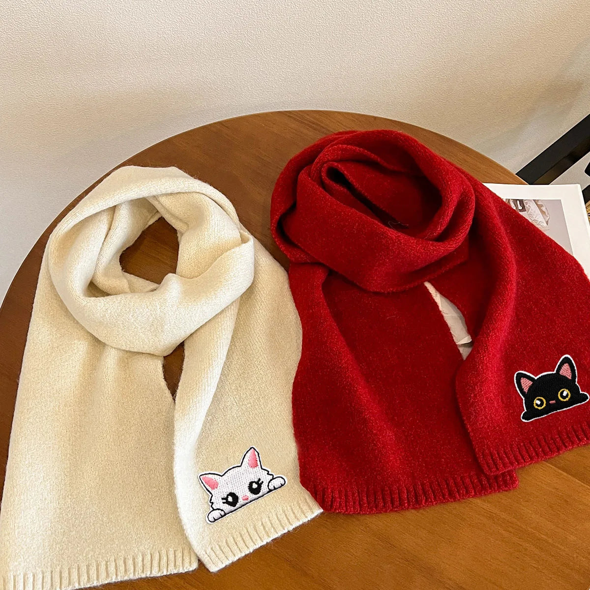 A beige knitted scarf with a cat embroidery patch lies next to a red knitted sweater.