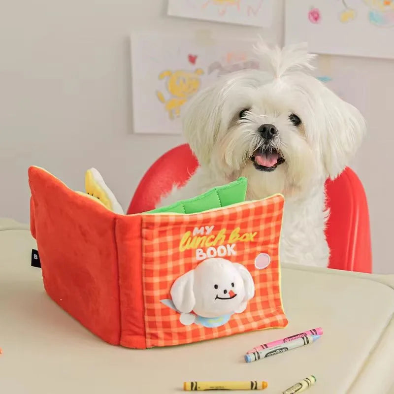 A white dog is looking at an orange fabric snuffle book toy with a white stuffed animal inside.