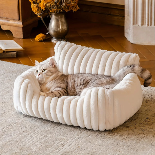Un chat gris et duveteux se repose dans un lit-canapé blanc à rayures sur un tapis clair.