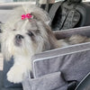 A fluffy white dog with pink hair accessory is resting in a gray portable pet car seat.