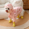 A small white dog wearing a pink cartoon printed raincoat with strawberry patterns and yellow boots.