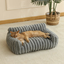 A fluffy orange cat is relaxing inside a gray, knitted sofa bed on a light beige carpet.