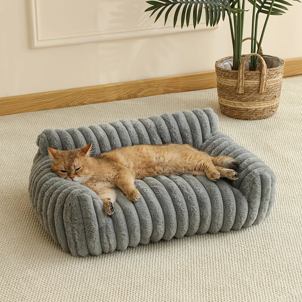 Un chat orange duveteux se détend dans un lit canapé gris tricoté sur un tapis beige clair.