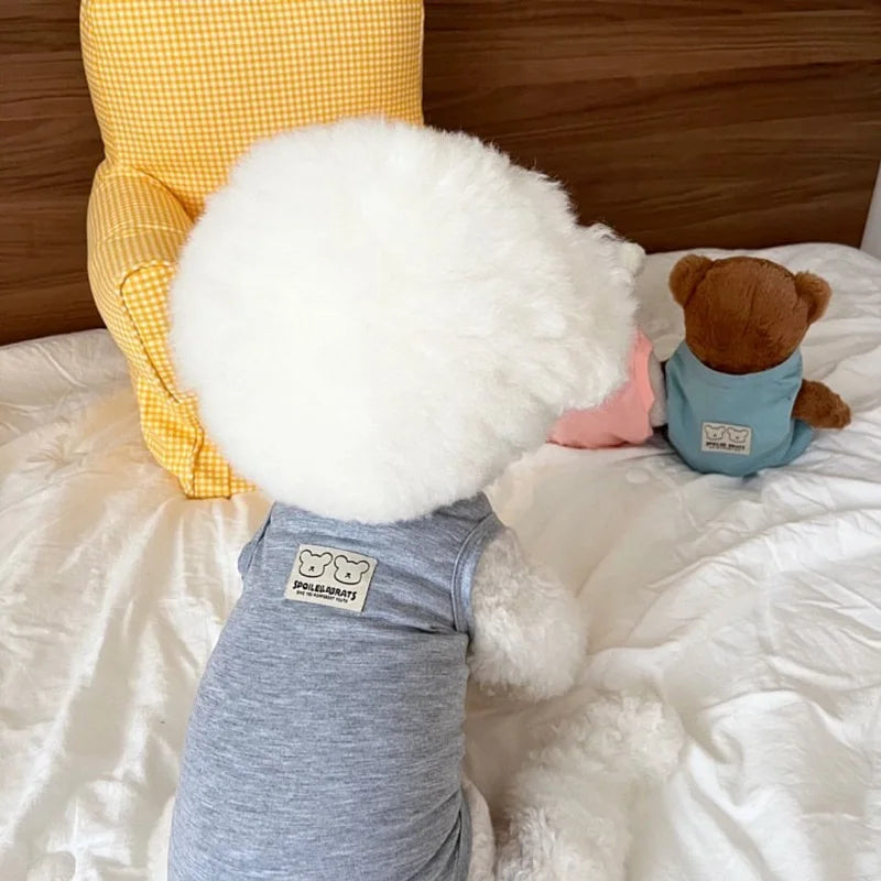 A fluffy white dog toy is lying on a white bed next to a blue vest and a brown teddy bear.