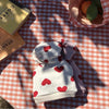 Sudadera de peluche con estampado de corazón blanco y rosa, doblada sobre un mantel a cuadros con luz del sol.