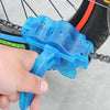 A hand holds a blue bike chain cleaner tool with a 3-sided brush system near a bicycle chain.