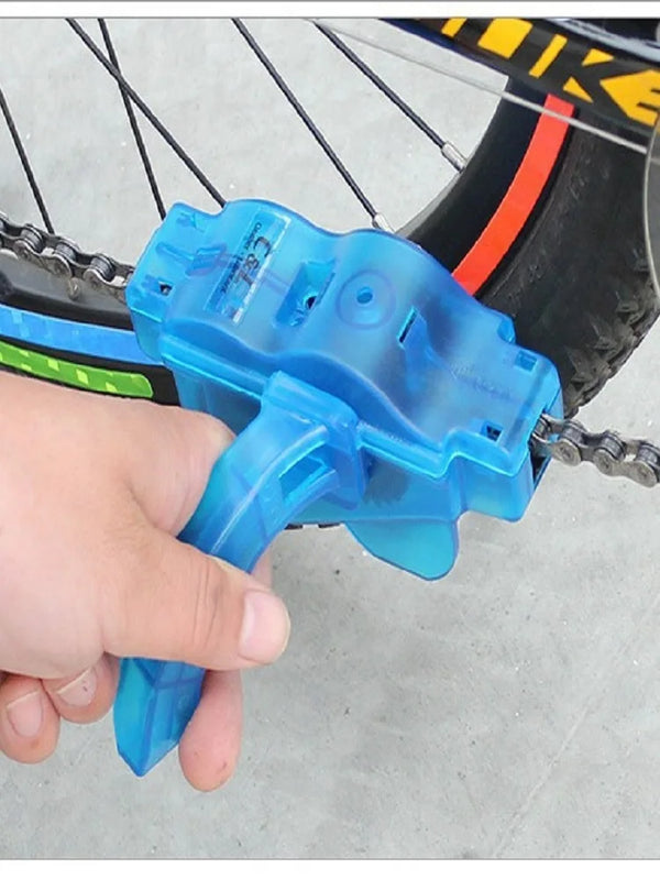 A hand holds a blue bike chain cleaner tool with a 3-sided brush system near a bicycle chain.