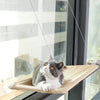 Grau-weiße Katze ruht in einem Holzfenster-Hängemattenbett, das an dünnen Drähten nahe einem Fenster hängt.
