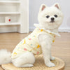A fluffy white dog wearing a yellow cotton vest with a lace collar and cartoon print, sitting on a wooden mat.