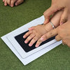 Hands are pressing a black handprint kit onto a white card on a green surface.