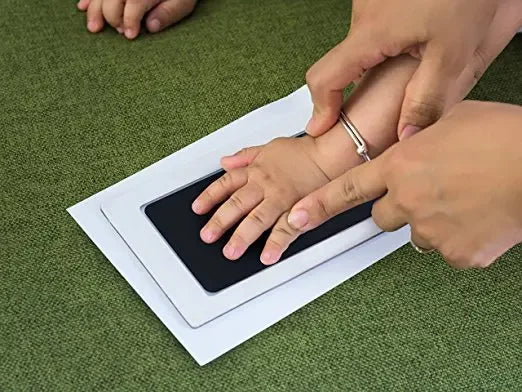 Hands are pressing a black handprint kit onto a white card on a green surface.