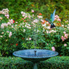 A solar-powered water fountain sprays water into a decorative bowl amidst lush green foliage and pink flowers.