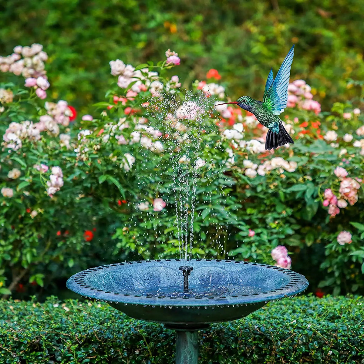 A solar-powered water fountain sprays water into a decorative bowl amidst lush green foliage and pink flowers.