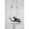 A black and white cat is resting in a macrame hammock pet bed hanging on a white door frame.