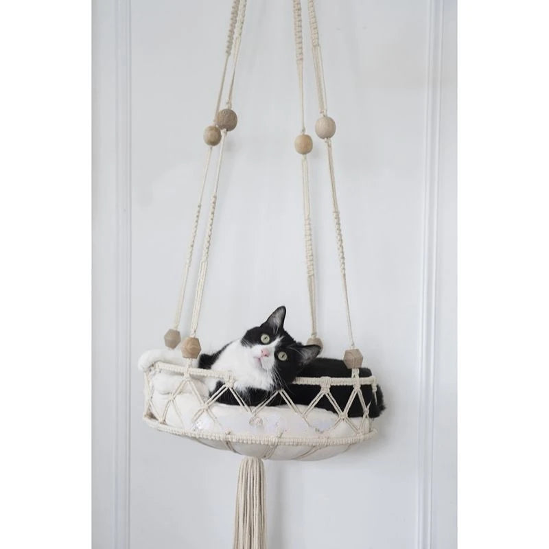 A black and white cat is resting in a macrame hammock pet bed hanging on a white door frame.