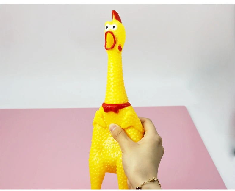 A hand is holding a yellow squeaky rubber chicken dog toy with a red beak against a pink background.