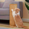 A fluffy orange and white cat is resting on a natural fiber scratch board placed next to a gray sofa.