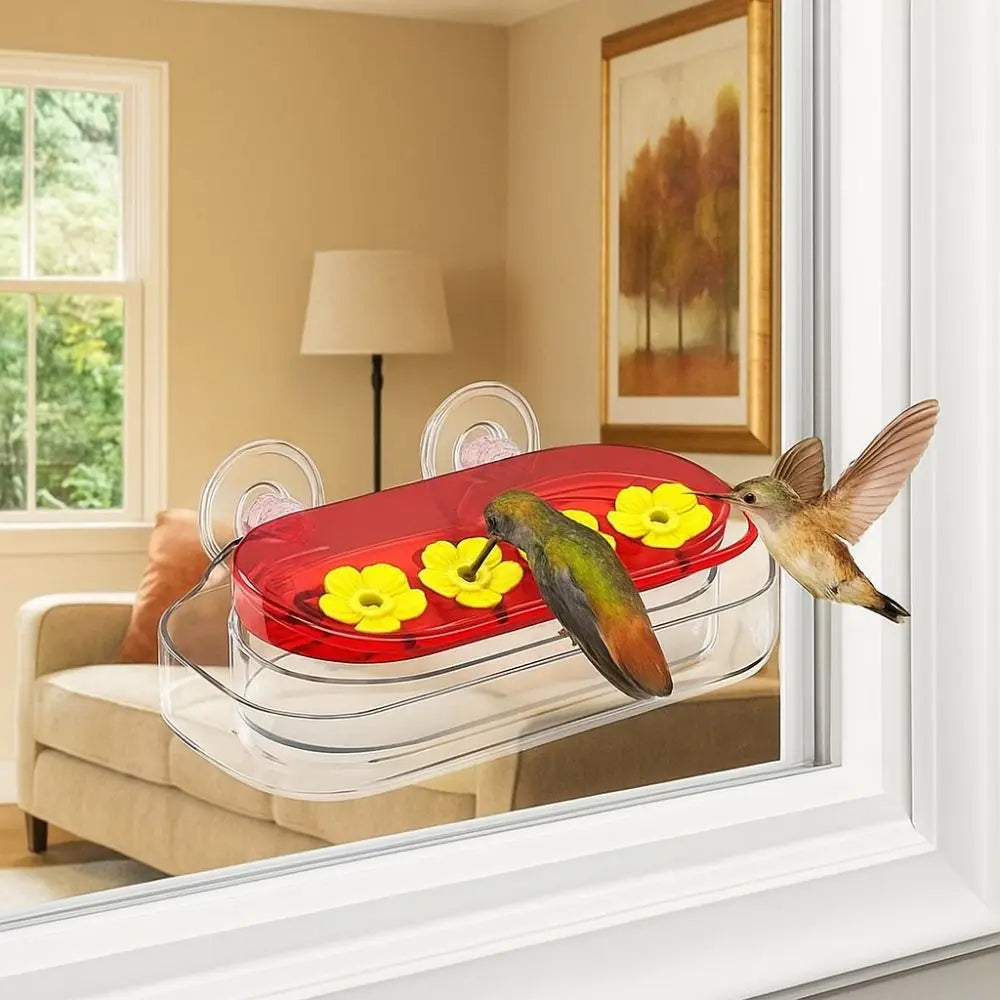 A clear plastic hummingbird feeder with a red reservoir and yellow flowers is placed on a windowsill.