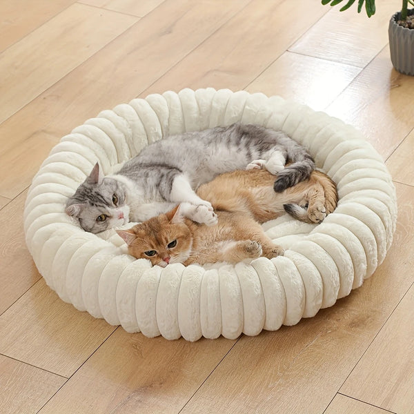 A round white pet bed with a plush bolster support is filled with three cats resting on a light wooden floor.