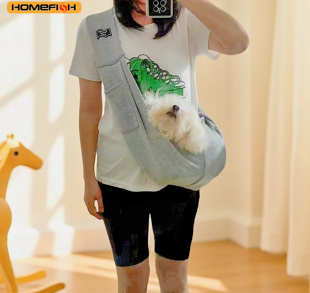 A person wearing a white t-shirt and dark shorts is holding an adjustable crossbody dog sling carrier with a fluffy white dog