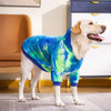 A light-colored dog wearing a blue and green tie-dye fleece hoodie standing on a floor.