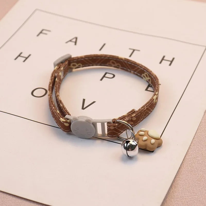 Brauner Lederhalsband für Haustiere mit silberner Sicherheitsglocke und Tan-Pfotenabdruck-Anhänger auf weißem Hintergrund.