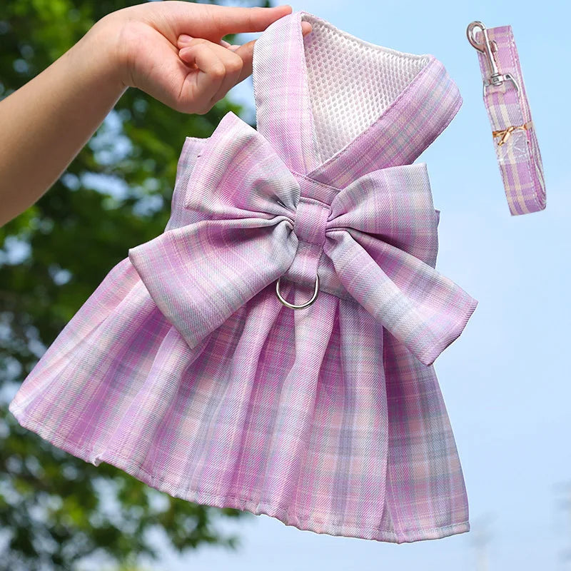 A pink plaid dog dress with a bow and harness is being held by a hand against a blue sky background.