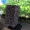 A rectangular block of black aquarium bio sponge filter media resting on a dark surface with green plants in the background.