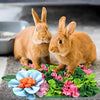 Two small brown rabbits standing on a green mat with pink and orange flowers