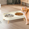 An orange cat sits next to an elevated cat bowl set featuring stainless steel food and water dishes.