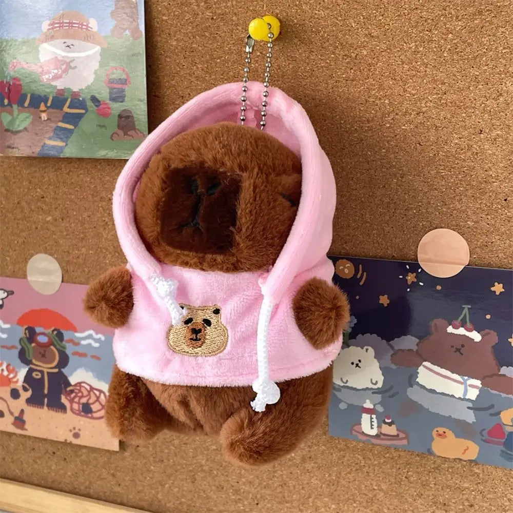 A brown capybara plush doll wearing a pink hoodie with white trim, displayed next to cards.