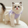 A fluffy white kitten wearing a lavender floral print dress with thin straps, sitting on a white surface.