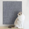 A white and gray cat is sitting in front of a dark gray textured scratching mat.
