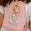 A clear, sculpted cat charm keychain with a gold-toned clasp attached to a pink backpack.